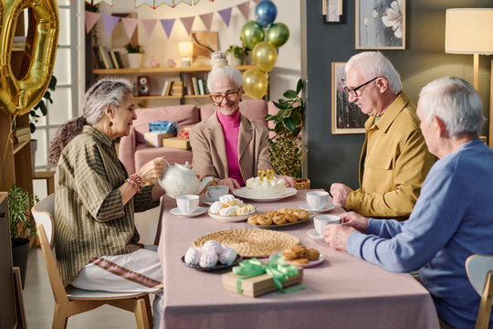 Group of senior Caucasian women and men sitting around table celebrating birthday, smiling and enjoying tea with cake and pastries, festive decorations and balloons in background