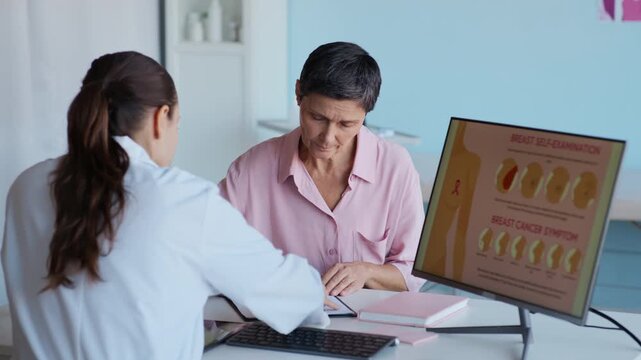 Medium shot of female oncologist in white coat guiding middle-aged patient filling medical form sitting at table with breast cancer illustration on computer screen in clinic