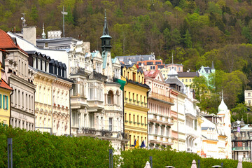 This is a vibrant view of Karlovy Vary, showing a row of colorful, ornate buildings nestled against a green, forested hillside. The architecture is a mix of styles
