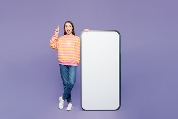 Full body young amazed woman wears yellow sweatshirt casual clothes big huge blank screen mobile cell phone smartphone with area point finger up isolated on plain purple background. Lifestyle concept.