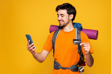 Young man carry backpack with stuff mat use mobile cell phone credit card isolated on plain yellow background. Tourist lead active lifestyle walk on spare time. Hiking trek rest travel trip concept.