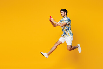 Full body side view young man he wearing blue beach t-shirt casual clothes jump high hold in hand use mobile cell phone isolated on plain yellow orange background studio portrait. Lifestyle concept.
