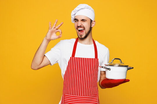 Young happy housewife housekeeper chef cook baker man wear red apron toque hat hold in hand saucepan show ok okay wink isolated on plain yellow orange background studio portrait. Cooking food concept.