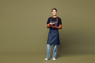 Full body young happy man barista bartender barman employee wear jeans apron black t-shirt work in coffee shop write down in notebook isolated on plain green background Small business startup concept