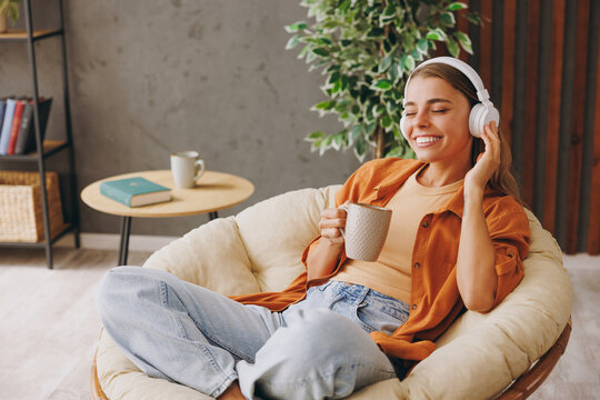 Young fun woman wear orange casual clothes listen music in headphones drink coffee sits in chair stay at home hotel flat rest relax spend free spare time in living room indoor. People lounge concept.