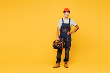 Full body young smiling happy employee laborer handyman man wear helmet overall hold bag with tools isolated on plain yellow background. Instruments for renovation apartment room. Repair home concept.