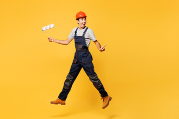Full body young employee laborer handyman man wear helmet overall jump high hold paint roller point aside isolated on plain yellow background Instrument renovation apartment room. Repair home concept