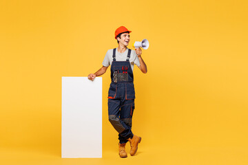 Full body young employee laborer handyman man wear helmet overall show big white wall scream in megaphone isolated on plain yellow background Instrument renovation apartment room. Repair home concept