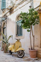 Fototapeten Roller Yellow vintage scooter in old city street of corfu, greece. Old stone house with wooden green shutters. Urban scene. Traditional greece  © Marjolein Hameleers