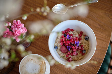oatmeal porridge with red frozen berries and violet flowers. A white bouquet of cherry branches in blossom period with wonderful white flowers and fresh green leaves in glass vase on a white table