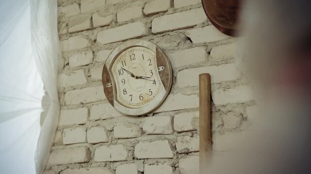 Old warped analog clock with distorted face hangs on white brick wall, ticking steadily as time passes, vintage style evokes surreal feeling of time slipping in rustic damaged indoor setting