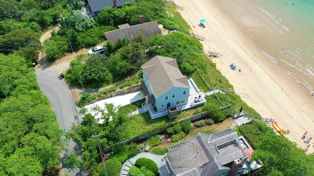 Aerial view of Cape Cod shoreline captures a beachfront home framed by dunes, beachgoers visible below, and turquoise waves glowing under warm summer light.