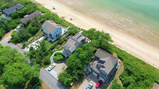 Cape Cod home overlooks sandy beach as families stroll below, turquoise water sparkles under bright sun, and a backdrop of trees completes the summer scene.