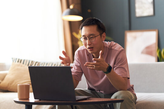 Young adult Asian man sitting on sofa gesturing with hands while having video call on laptop, wearing glasses and smartwatch, appearing engaged in online conversation at home
