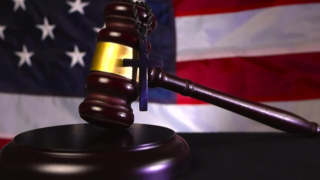 Wooden court justice gavel resting on a wooden dark desk with a holy christianity cross hanging on the bronze decorations and a American United states flag in the bokeh blurry background.