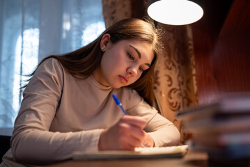A young girl is focused on doing her homework. A student is sitting at a desk and writing in a notebook, concentrating on her studies