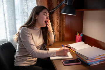 A female student yawning from fatigue while studying. A young girl, exhausted from studying, is sitting at a desk and yawning, covering her mouth with her hand