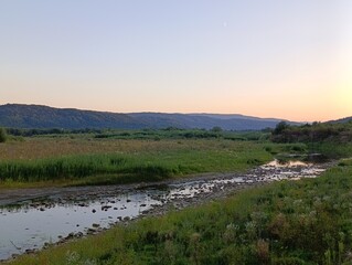 Sunset over a serene river landscape with rolling hills and lush greenery at twilight