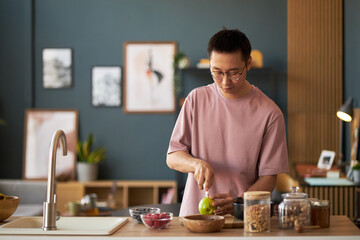 Young adult Asian man preparing food in modern kitchen, slicing green apple on wooden cutting board, surrounded by glass jars and bowls with ingredients, wearing eyeglasses