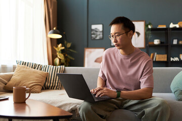 Asian young adult man sitting on sofa using laptop, focusing on typing while working or studying in modern living room, coffee mug on table in foreground, natural daylight streaming in