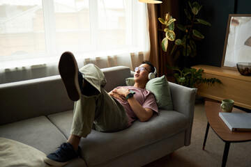 Asian young adult man reclining on sofa with headphones, relaxing with eyes closed and hands resting on chest, enjoying leisure time in modern living room with natural light