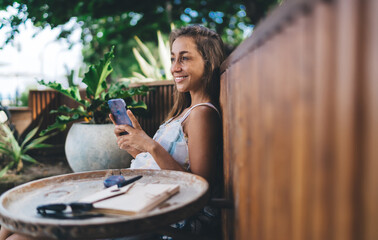 Smiling woman gazing sideways while holding smartphone, embracing online connection with joyful ease, showcasing tech as emotional extension of modern lifestyle in digital leisure environment.