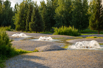 three water geyser fountains in modern park garden, lush greenery and stone hills pathways. The warm light of sunset enhances the calm ambiance of the area.