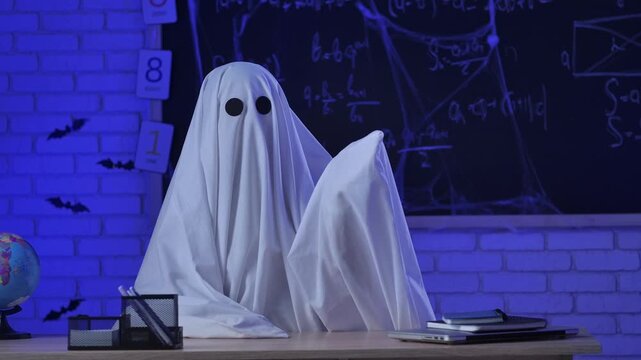 A person in a white ghost sheet sits at a table in a blue-lit room and waves hello