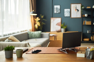 Modern home office desk featuring open laptop, smartphone, potted plants on wooden surface with blurred living room and framed wall art in background, daylight streaming through window
