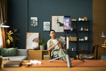 Young adult Asian man sitting in modern living room holding notebook and pen, looking at smartphone...