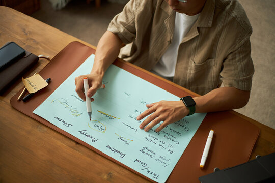 Young adult Asian man sitting at desk writing on large sheet of paper with marker, planning project tasks and priorities, using smartwatch and digital tablet for organization