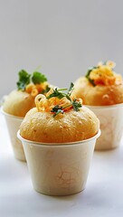 Three fluffy, light-colored baked goods in small paper cups, topped with a sprinkle of cheese and parsley, sit on a white surface against a muted background