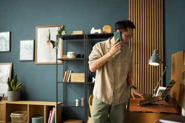 Asian young adult man standing at desk talking on smartphone while using laptop in modern workspace, looking focused and engaged in multitasking with technology