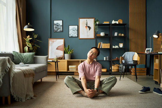 Young adult Asian man sitting cross legged on carpeted floor in modern living room, stretching legs and holding feet with both hands looking slightly upward with neutral expression