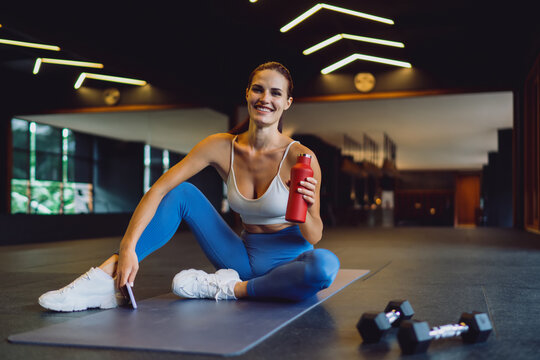 Smiling fit woman in activewear sits on yoga mat after workout, holding red water bottle and smartphone, symbolizing healthy lifestyle, digital balance, and body-mind connection.