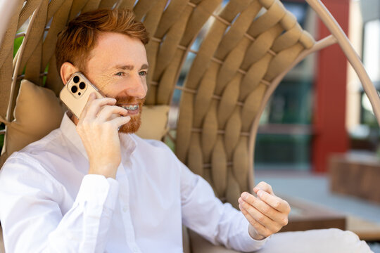 Red-haired businessman relaxing in lounge chair outside talking on smartphone - Powered by Adobe