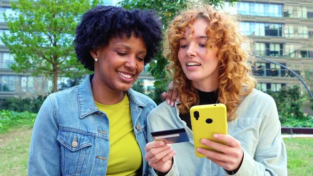  Cheerful African American and Caucasian friends stand in a city park, holding colorful smartphones and a credit card, celebrating cashless convenience, shared shopping success, and digital lifestyle