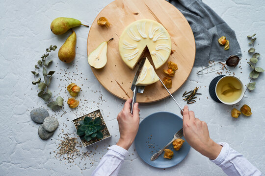 Caucasian young adult woman cutting cheesecake with knife and serving slice onto plate, surrounded by pears, tea, succulents, and decorative elements on light textured surface