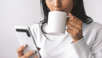 Young woman in a white sweater drinks from a clean white coffee mug while looking at her smartphone.