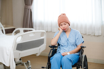 A female patient wearing a beanie and hospital gown sits in a wheelchair in a hospital room beside a bed, looking thoughtful and somewhat somber. The scene conveys rest,