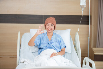 A female cancer patient wearing a knit hat is sitting up in a hospital bed, smiling and making an "OK" hand gesture. She appears positive and hopeful during her recovery, 