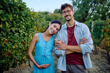 Naklejka premium Smiling young pregnant woman leaning on husband's shoulder, standing together in a vineyard. Celebrating new life outdoors