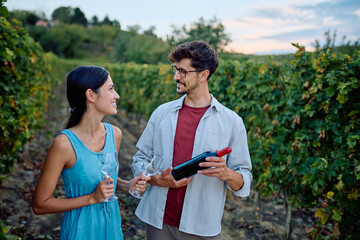 Smiling young couple holding wine bottle and glasses, enjoying a romantic date outdoors in a beautiful vineyard setting