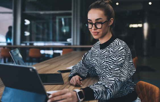 Focused freelancer woman in glasses interacting with tablet, managing data across devices, reflecting agility, productivity and synchronized digital performance in creative environment.