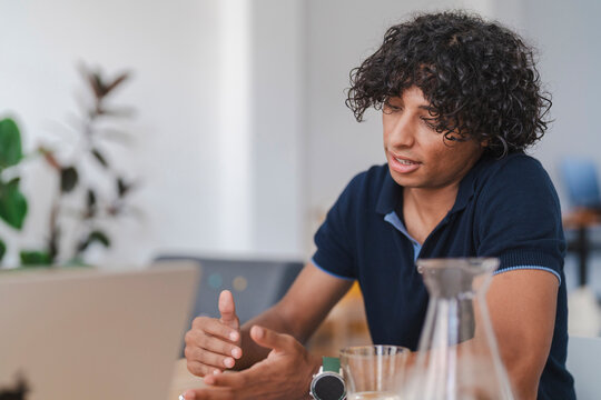 A young man with curly hair sits at a table, looking at a laptop. He gestures with his hands while speaking, engaged in a virtual meeting or discussion.