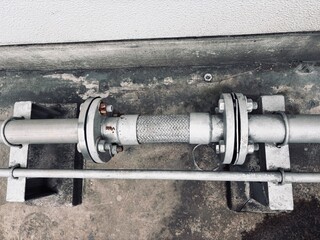 Close-up of a braided stainless steel flexible metal hose/joint connecting industrial pipes with bolted flanges, showing the metallic texture and surrounding overgrown weeds.