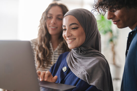 Three diverse young adults are smiling and collaborating around a laptop. A woman in a hijab interacts with the screen, sharing content with her peers.