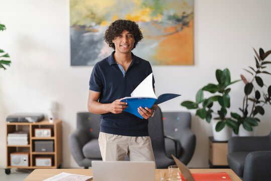 A cheerful young man with curly hair stands in a contemporary workspace. He holds an open blue notebook, smiling confidently at the viewer, ready for his tasks.