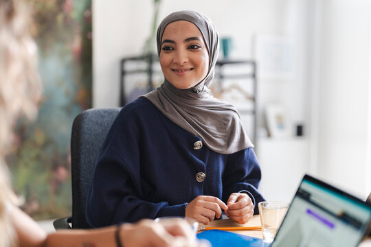 A smiling young Muslim woman in a hijab sits at a table, engaged in a conversation. She is in an office setting with a laptop and other items on the desk.