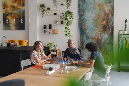 Diverse young professionals collaborate on laptops at a modern office table. Two women and one man engage in a productive discussion during their workday.